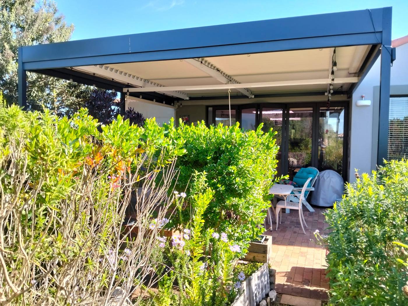 Charcoal louvre roof over a brick-paved patio with a cream drop blind lowered on the side