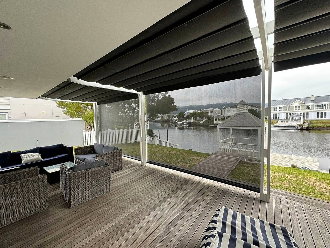 Mesh drop screens closing the sides of a timber deck under a charcoal retractable canopy, facing a canal