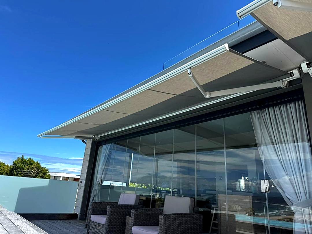 Grey retractable awning on folding arms shading a terrace with glass doors and a glass balustrade